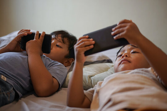 Two children lying on bed using smartphones, holding screens close to face. Indoor lifestyle and screen time concept, selective focus