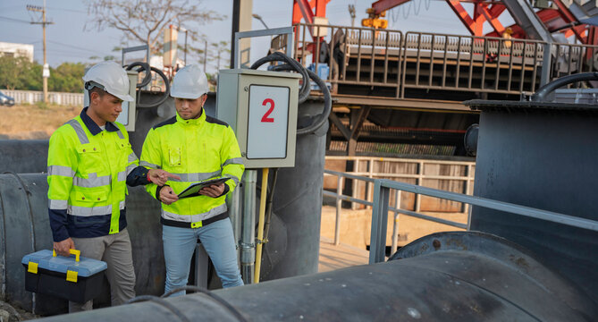 Professional Asian engineers inspecting industrial water pump machinery. Two technicians checking maintenance records on a clipboard at an outdoor construction site factory facility.