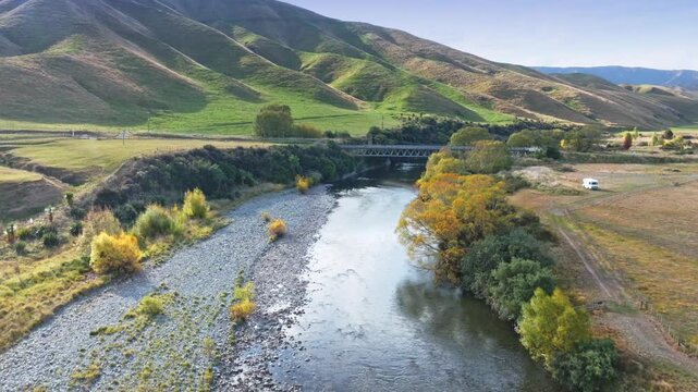 Autumn colors frame a wide river flowing through a valley in Manawatu-Whanganui, New Zealand. The landscape features rolling hills and lush green fields under a clear blue sky.