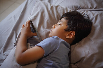Young boy lying in bed using smartphone while resting on pillow. Screen time and indoor lifestyle concept © tuan12
