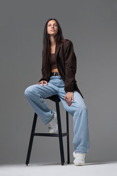 Fashion model in brown blazer and wide jeans posing on high chair against gray wall