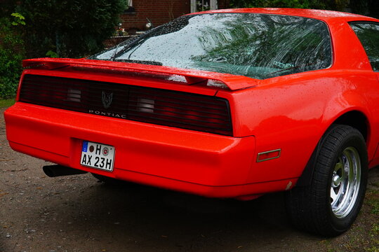 An orange red Pontiac Firebird, rear end featuring the Pontiac logo and crest. Raindrops on the red paint in Hanover, Germany.