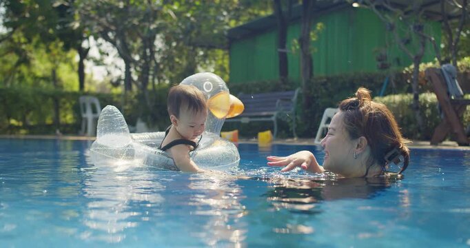 An Asian mother lovingly supervises her toddler in a duck floatie, fostering water confidence and enjoying a bonding moment during a family swim in a pool.