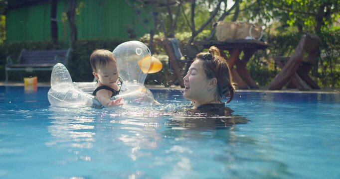 An Asian mother teaches her toddler to swim in a resort pool, fostering early development and family bonding during a fun summer vacation activity.