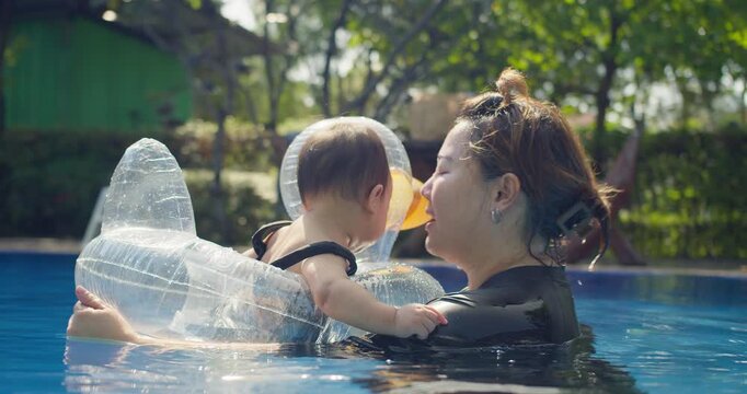 An affectionate Asian mother kisses her toddler in a pool, fostering a close bond while introducing the child to swimming in a safety flotation device.