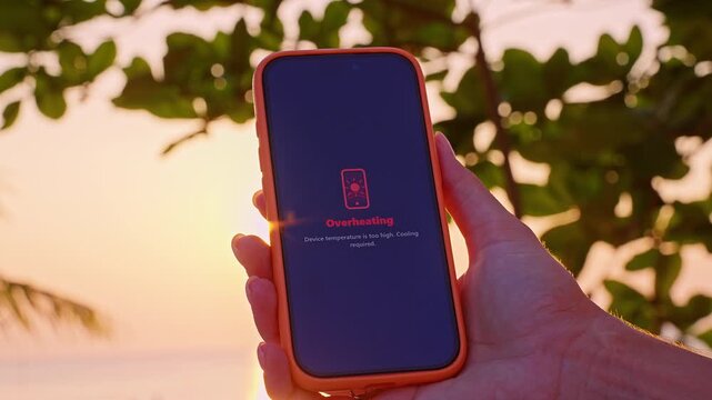 Smartphone Screen Showing Overheating Warning Message in Hand at Sunset with Tropical Background, Device Temperature Concept