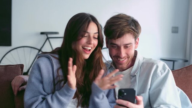 A happy young couple sits on a comfortable sofa at home, sharing an exciting moment as they react with surprise and joy to something seen on a smartphone screen.