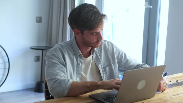 A focused young man diligently works on his laptop, typing and taking notes, embracing the modern remote work lifestyle from his comfortable home. He is engaged in productive tasks managing