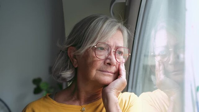 An elderly woman with grey hair and glasses looks out her window with a thoughtful expression. She appears to be contemplating life or reminiscing, creating a serene and reflective mood indoors.