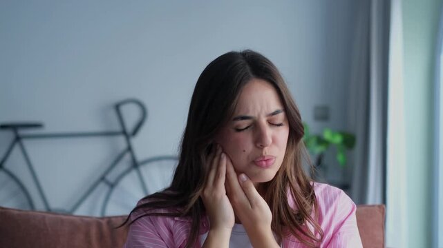 A young woman sitting at home experiences sharp toothache or jaw pain. She touches her cheek and winces, showing discomfort and suffering from a dental problem, seeking relief.