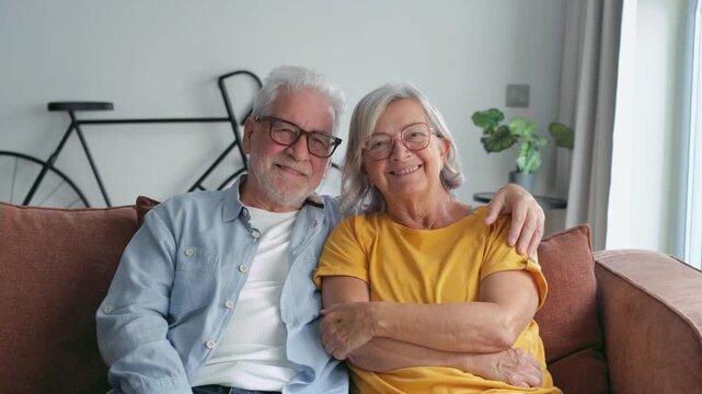 A happy senior couple sits comfortably on their couch at home, embracing and sharing a loving smile. They radiate contentment and affection in their golden years, enjoying a peaceful domestic life