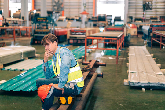 Stressed factory worker in yellow safety vest sitting on steel beam at industrial warehouse, hand near mouth looking worried and thinking, blue collar burnout work pressure mental health challenge