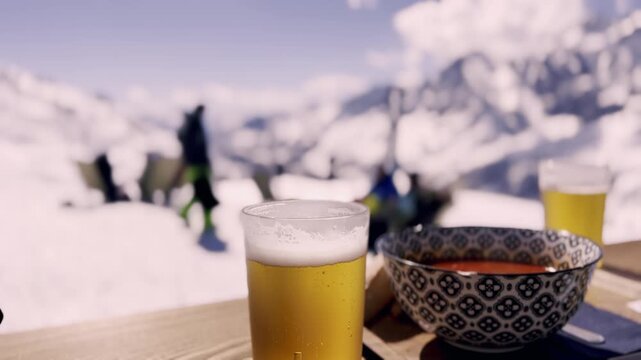 Refreshing glass of beer and a warm bowl of soup resting on a wooden table at a ski resort, with skiers and snowy mountain peaks creating a beautiful blurred background on a sunny day