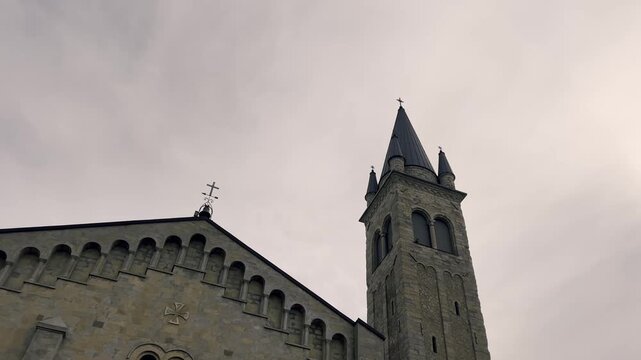 Low-angle view of the romanesque stone facade and the tall belfry of a medieval christian church, with a somber and imposing atmosphere under an overcast and cloudy sky