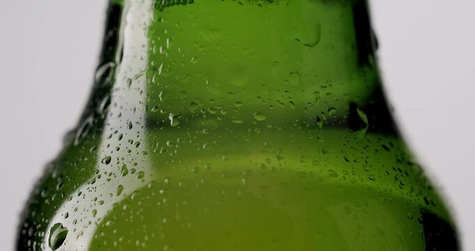 Macro view of a chilled green glass beer bottle covered in fresh water drops and condensation, slowly rotating in place against a clean, isolated white studio background.