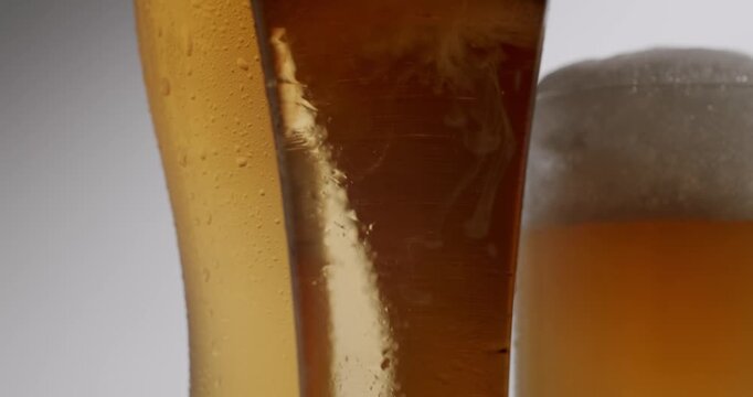 Refreshing amber beer pouring into a glass in slow motion creating white foam, bubbles, and condensation. Two glasses of cold lager beer isolated on a plain studio white background