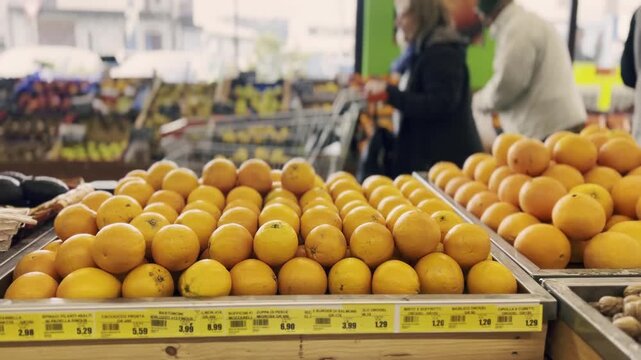 Customers shopping for fresh produce in the fruit and vegetable section of a supermarket, with a foreground focus on ripe oranges and walnuts displayed in wooden crates with price tags