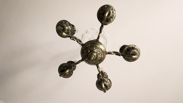 Low angle view of an elegant antique bronze chandelier with six arms swaying gently from a white ceiling with a decorative rosette, creating moving shadows and light reflections on the surface