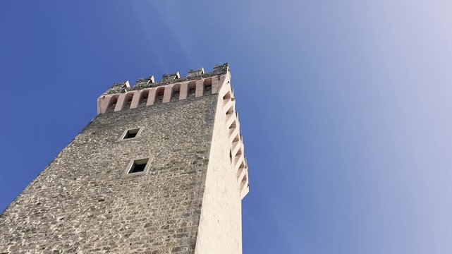 Low-angle perspective of an ancient medieval stone tower with battlements, standing tall against a bright and clear blue sky on a sunny day, showcasing historical architecture and fortitude