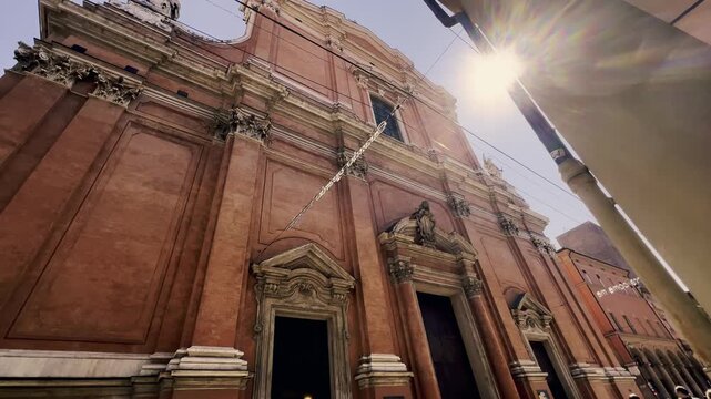 Low angle view of the ornate baroque facade of the Oratory of San Filippo Neri in Bologna, Italy, with the sun flaring brightly and the Garisenda Tower visible in the background