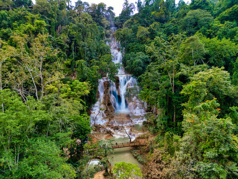 Aerial view of Kuang Si Waterfall with cascading turquoise water tiers surrounded by lush tropical jungle and a wooden bridge in Luang Prabang, Luang Prabang Province, Laos.