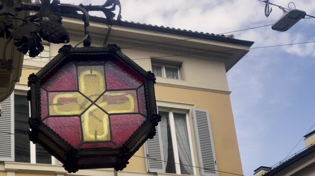 Antique wrought iron lantern with a red and yellow glass cross signaling a historic pharmacy in an old European city, swaying gently with a traditional building facade in the background