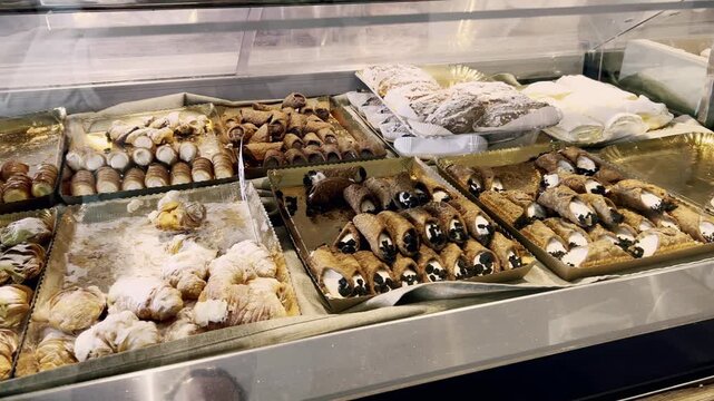Wide assortment of traditional sicilian desserts, including fresh cannoli filled with ricotta cream and chocolate chips, displayed on trays in a refrigerated pastry shop counter