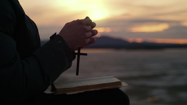 The hands of a man with a Christian cross reading the Bible on the shore of a lake at sunset. A man studies the word of God in search of truth and spirituality.
