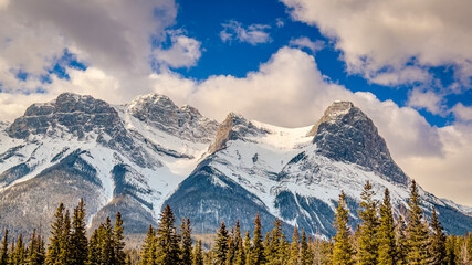 Snow‑covered mountain range framed by evergreen forest under a bright blue, partly cloudy sky — dramatic alpine landscape for travel, nature, and outdoor themes © Greg