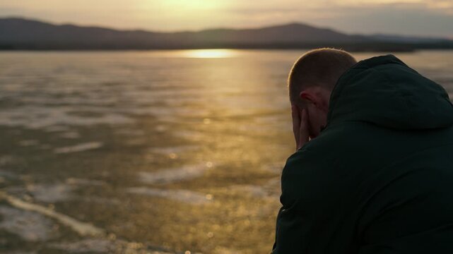 A sad male sits alone on the shore of a lake at sunset. Loneliness with oneself, despondency. The restless psychological state of a person.