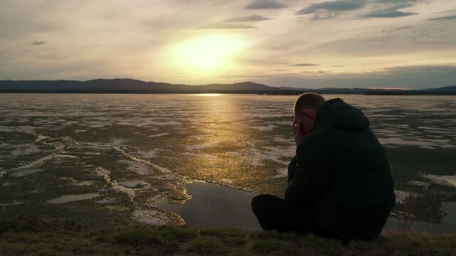 A sad male sits alone on the shore of a lake at sunset. Loneliness with oneself, despondency. The restless psychological state of a person.
