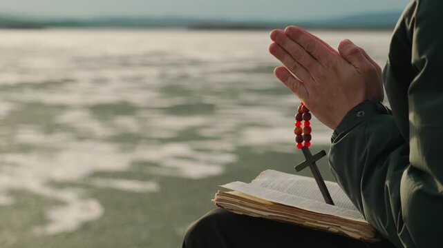 The hands of a man with a Christian cross reading the Bible on the shore of an icy lake. A man alone studies the word of God in search of truth and spirituality.