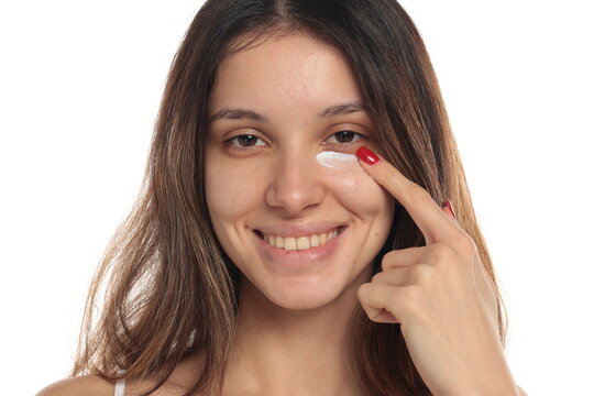 Studio shot of a smiling brunette woman applying eye cream under her eye with finger, portrait, headshot, on a white background