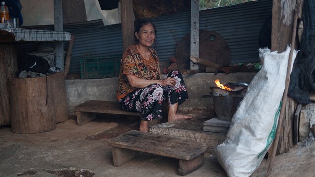 Elderly woman sitting on wooden bench preparing food on a fire in a rustic home in southeast asia