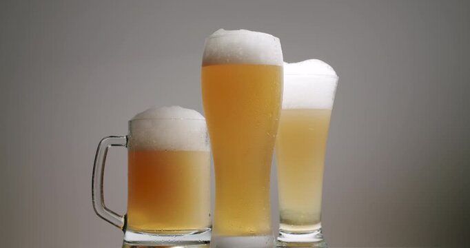 Three glasses of cold, fresh beer with a thick white foam head slowly rotating on a white background. Different types of beer glasses filling up with light ale and overflowing with thick white foam