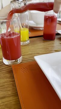 Vertical 9:16 POV of a hand pouring vibrant red juice into a glass during a hotel breakfast, followed by a close-up of a fresh omelet, bacon, and broccoli on a wooden table. 