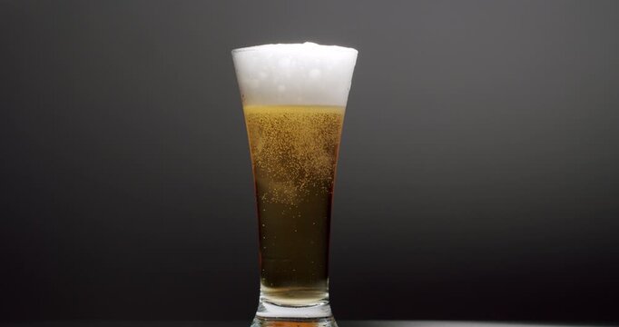 Cold light beer in a glass with bubbles rising to the top, creating a white foam head that slowly overflows on a dark studio background, showing the refreshing quality of the beverage