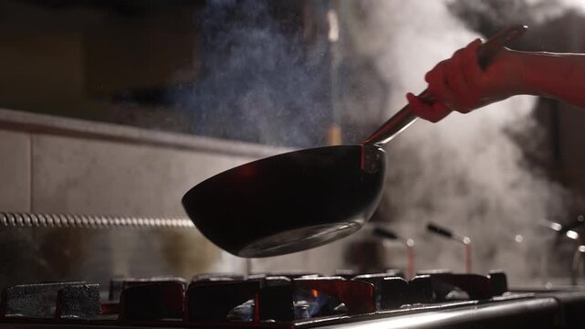 Chef in a restaurant kitchen flambeing food in a wok over a hot stove with lots of smoke, fa01
