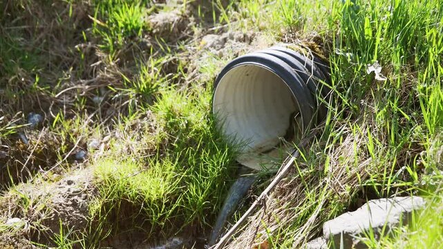 Black corrugated drain pipe discharging clear water into grassy natural landscape under sunlight