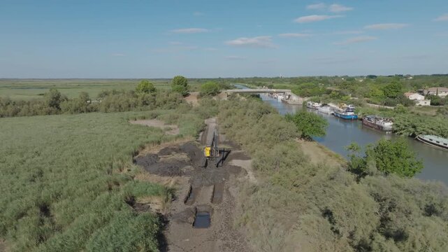 Aerial view of Camargue canal with yellow excavator digging dark mud on the bank near moored boats and a bridge under bright blue sky