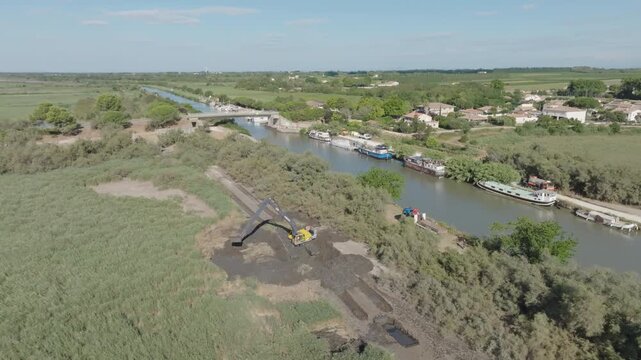Aerial view of Camargue canal with yellow excavator digging dark mud on the bank near moored boats bridge and green fields under bright blue sky