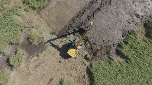 Top down aerial view of Camargue wetland with yellow excavator positioned on large dark mud pile beside green marsh vegetation