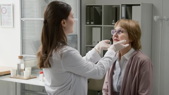 Dolly in shot of young adult female physician wearing protective gloves palpating lymph nodes while examining elderly woman during routine physical exam at clinic