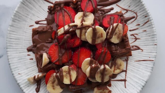 Close-up top down zoom in view massive, high-calorie waffle topped with strawberries, bananas, and chocolate at an outdoor cafe. A delicious and indulgent big portion sweet treat