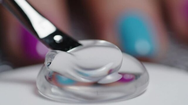 Close-up of clear liquid being moved and manipulated with a metallic tool on a white surface with blurred colorful background