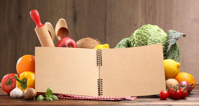 Open recipe book, kitchen utensils and fresh products on wooden table, closeup. Space for text