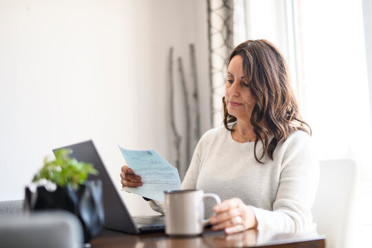 Senior woman at home kitchen table feeling stressed and confused while looking at multiple bills working on her laptop