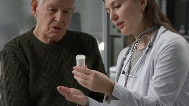 Tilt up shot of young adult female doctor explaining elderly man how to take medication while holding bottle of pills and discussing treatment options with patient in medical office