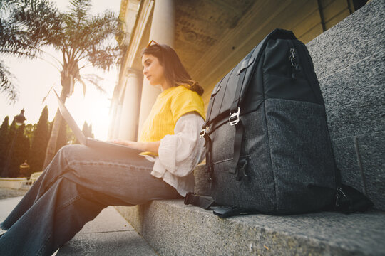 Young woman in yellow with backpack and laptop outdoors in an urban setting at sunset. Modern lifestyle and remote work concept.