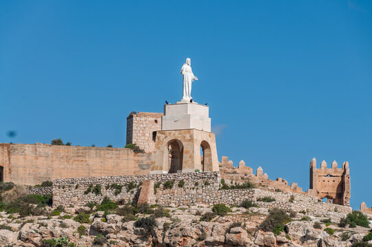Statue of Christ overlooking the landscape, proudly standing on a stone pedestal integrated with ancient fortifications remnants atop a rocky hill under a clear blue sky